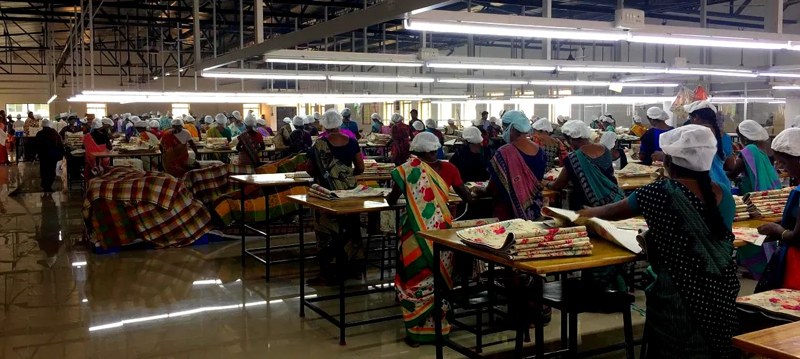 Production floor in India: rows of work tables where staff sort and pack printed textiles, under an open steel structure with strip lighting.